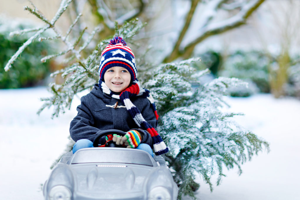Funny little smiling kid boy driving toy car with Christmas tree. Happy child in winter fashion clothes bringing hewed xmas tree from snowy forest. Family christmas tradition, holiday