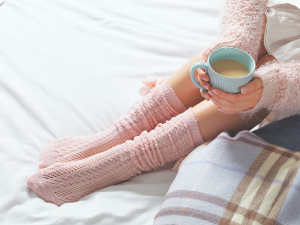 Woman relaxing at cozy home atmosphere on the bed. Young woman with cup of cocoa or coffee in hands and cookies enjoying comfort. Soft light and comfy lifestyle concept.